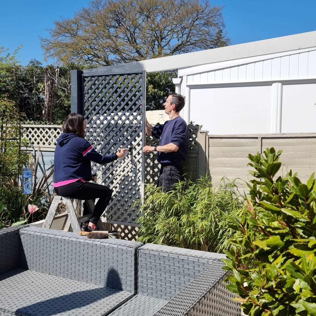 Two people are installing a lattice fence panel in a sunny garden. One stands and holds the panel, while the other kneels on a stepladder; both work together beside a patio seating area and green plants during their April monthly gardening.