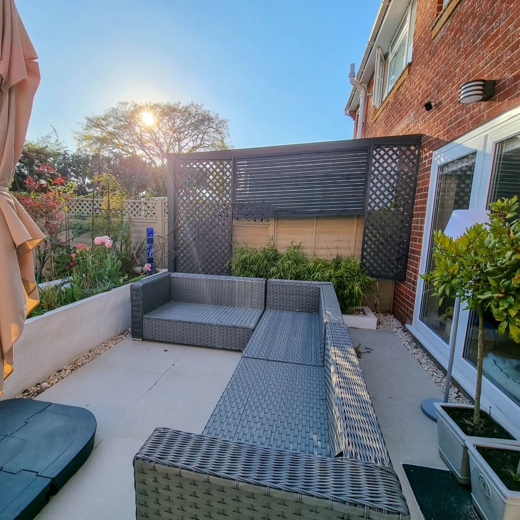 A modern patio with a grey wicker L-shaped sofa, light stone flooring, tall fence panels, lush garden plants, and a sun umbrella. The sun is low in the April sky, casting warm light over the inviting outdoor space.