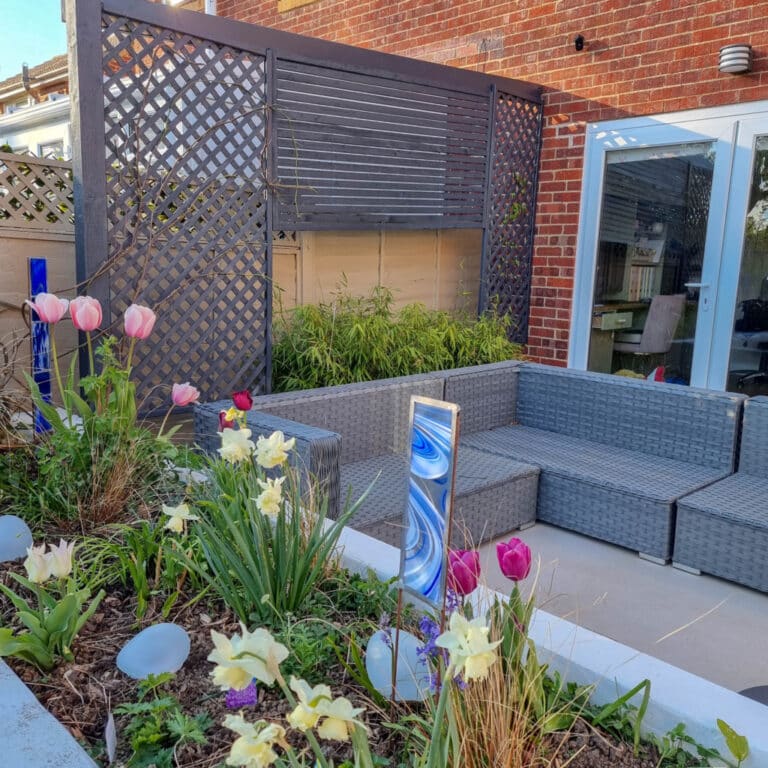 A modern patio with grey wicker seating, a trellis screen, and glass doors. The foreground garden blooms with April tulips and daffodils, accented by a decorative vertical garden mirror.