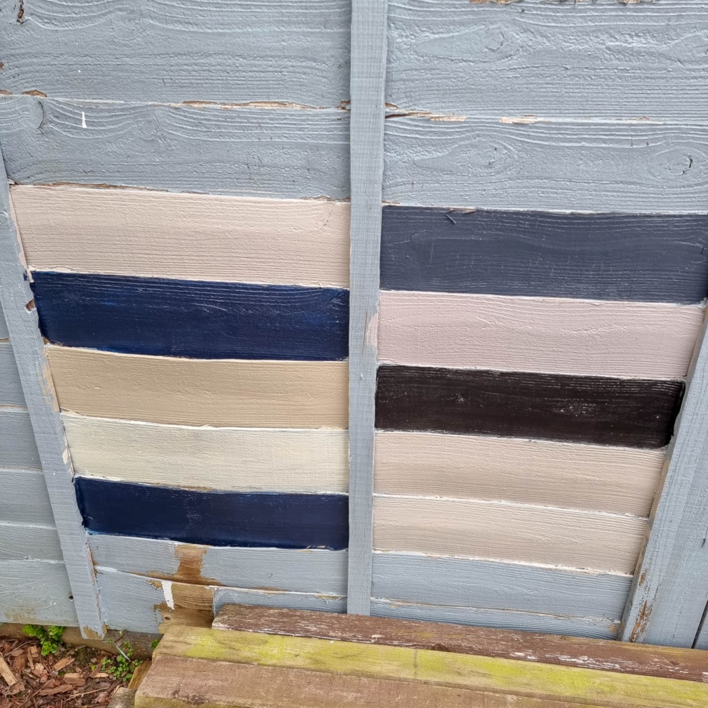 A wooden fence in a garden displays four horizontal paint swatches on each side of a vertical plank, featuring shades of navy, cream, beige, brown, and black. April leaves and wood are visible at the bottom.