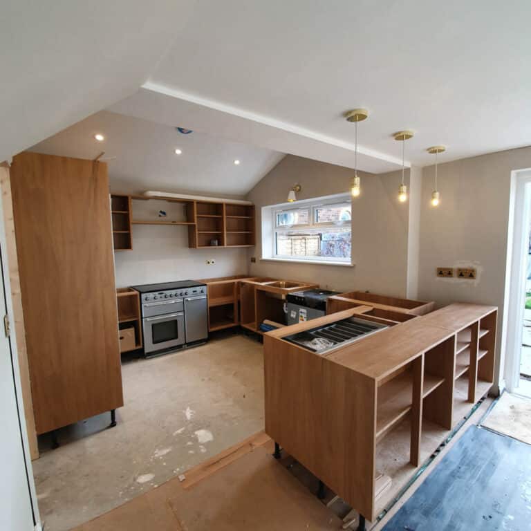A modern kitchen under construction with wooden cupboards, an unfinished island, open shelving, a large cooker, Creative Cables pendant lights, and an open window showing the outdoor garden.