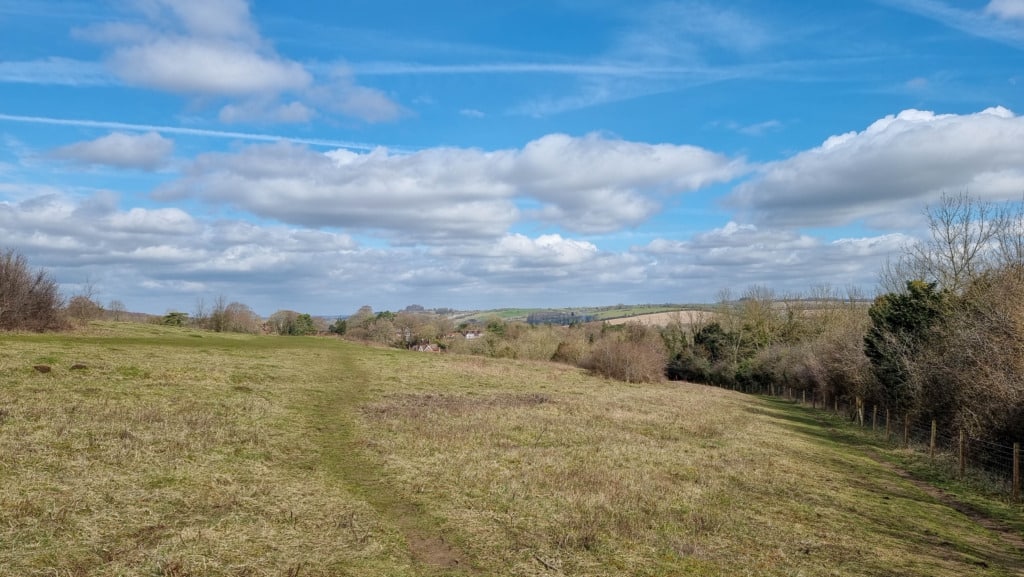 A grassy path offers a small walk through an open field bordered by trees under a blue sky with scattered clouds. Shawford Down’s rolling hills and distant houses are visible in the background.