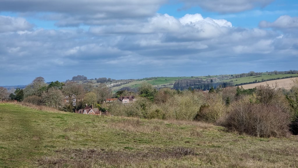A rural landscape with rolling hills, scattered trees, and a cluster of houses with red roofs partially hidden among the greenery under a cloudy blue sky—perfect for a small walk near Shawford Down.