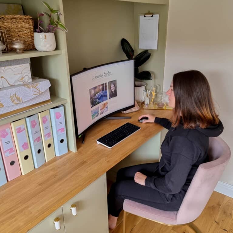 A woman in comfortable clothing sits at a wooden desk, using a computer with a large curved monitor. Shelves with boxes, files, plants, and decor are in the background. The workspace looks tidy and organised.