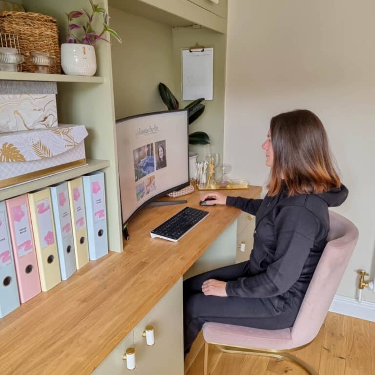 A woman in comfortable loungewear sits at a wooden desk in a home office, working on a computer. Shelves with boxes, files, and potted plants are in the background. The room has a calm, organised atmosphere.