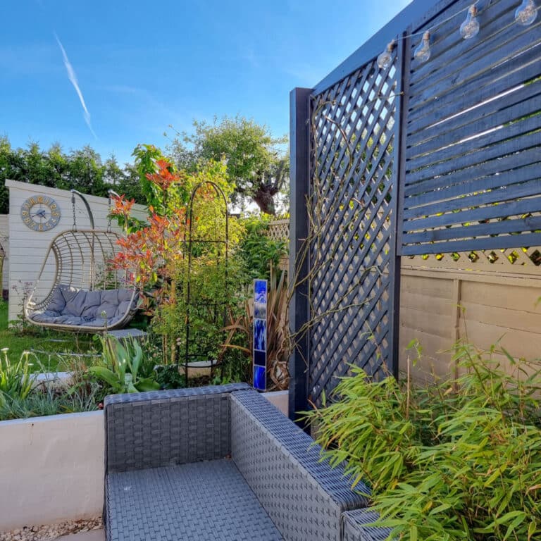 This month, enjoy a sunny May garden with green plants, a grey wicker outdoor sofa, a hanging egg chair, wooden lattice fence panels, fairy lights, and a blue garden ornament under the clear blue sky.