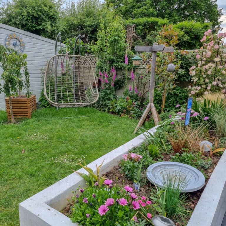 A lush June garden with a white raised flower bed full of colourful flowers, a wooden bird feeder, a hanging chair, and a garden shed with a decorative clock. Trees and flowering plants surround the green lawn, perfect for gardening enthusiasts.
