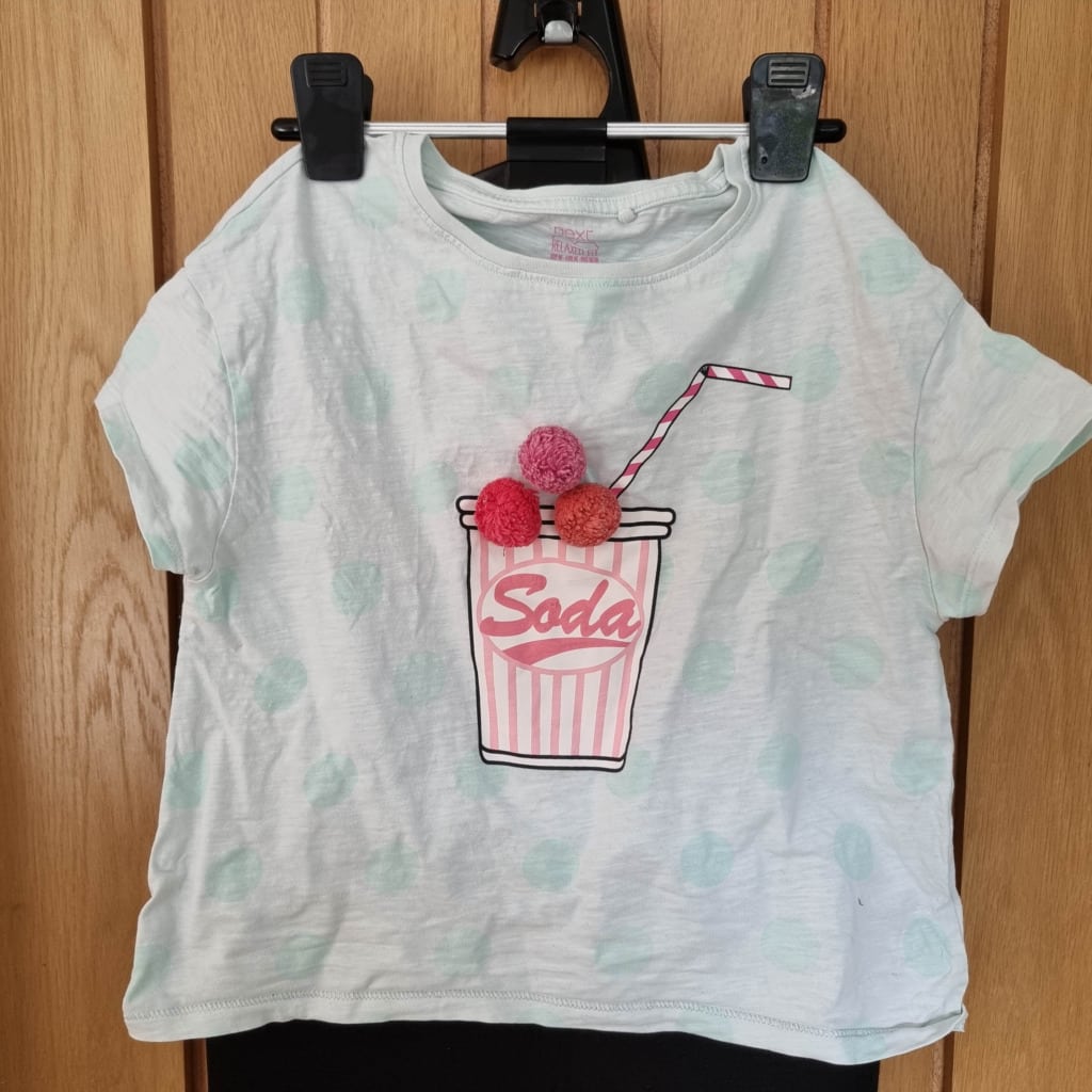 A white children's t-shirt with light green polka dots features a graphic of a pink striped fizzy drink cup and three red pom-poms, hanging on a door. Perfect for trying out your Handheld Garment Steamer for smooth, crease-free style.