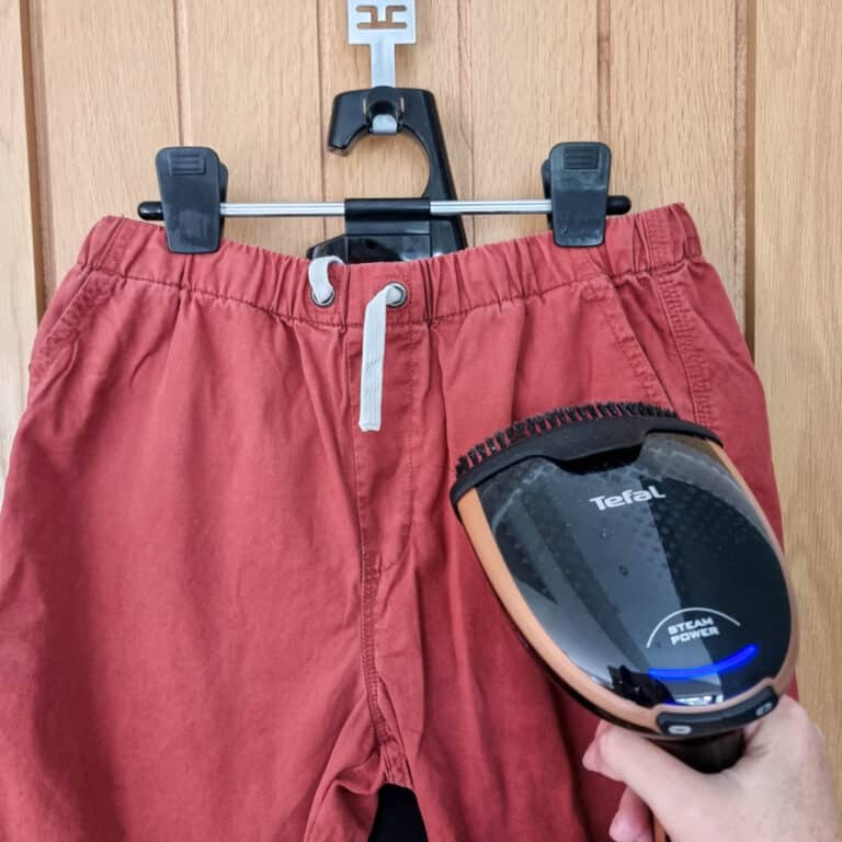 A person uses the Tefal DT9100 Handheld Garment Steamer to remove creases from a pair of red shorts hanging on a black plastic hanger against a wooden door.