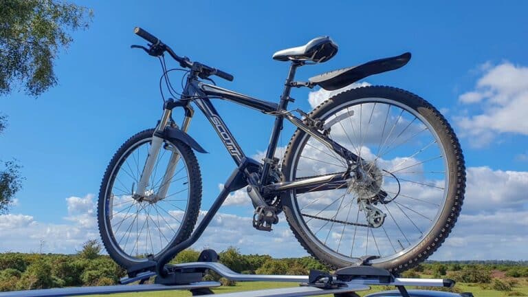 A black and silver mountain bike, perfect for cycling even in winter with the right tips, is mounted on a car roof rack under a bright blue sky with fluffy white clouds and a green landscape in the background.