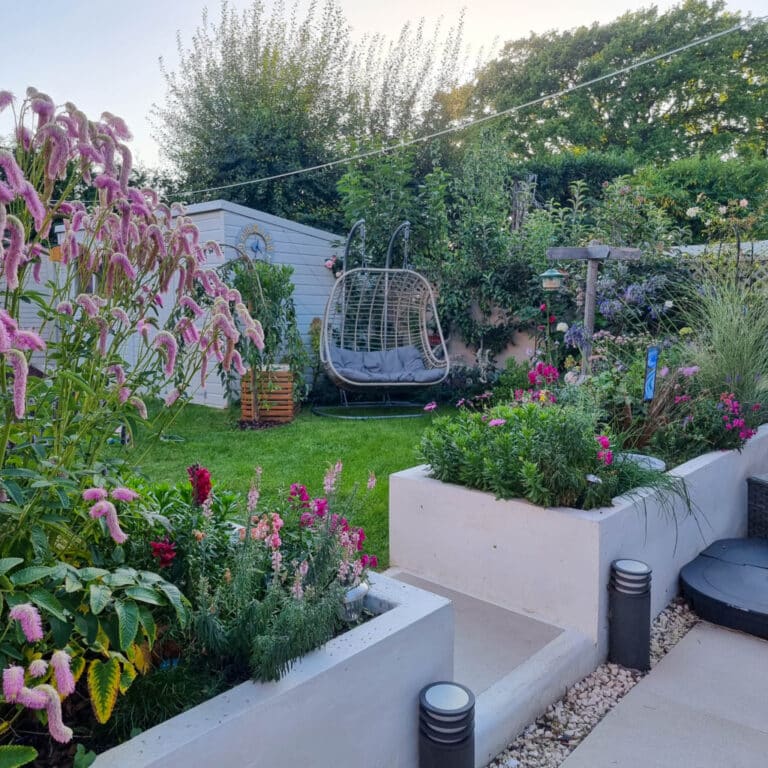 A lush garden this month with colourful flowers, green grass, a grey egg-shaped swing chair, white raised planters, and a modern patio. Shrubs and trees provide privacy in the background at the end of the summer.