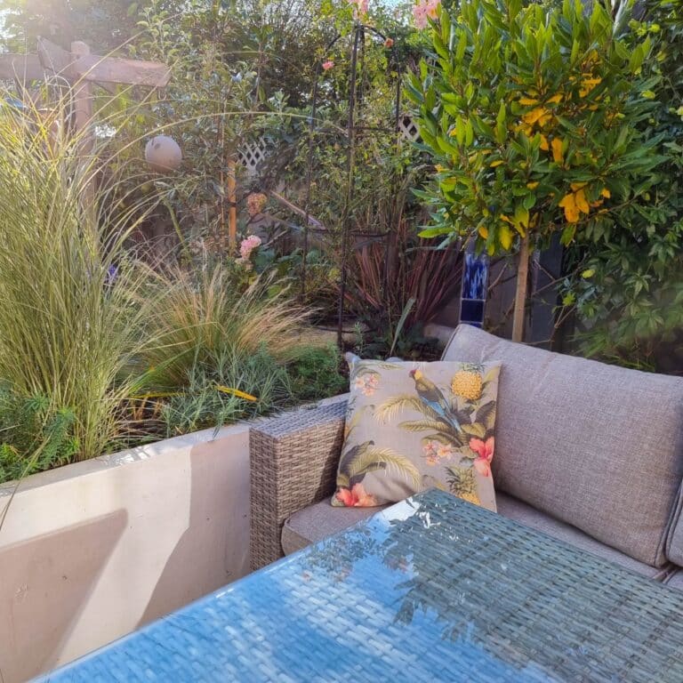 A cosy outdoor patio with rattan garden furniture, including a cushioned sofa, a tropical-print cushion, and a glass-topped table. Lush plants and flowers fill the background as sunlight casts soft shadows across the scene.