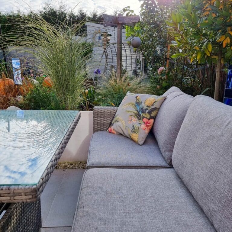 A cosy outdoor patio with grey rattan garden furniture, including a wicker sofa, glass-topped table, and a cushion with bird and flower designs. Lush greenery and garden decorations create a relaxing atmosphere in the background.