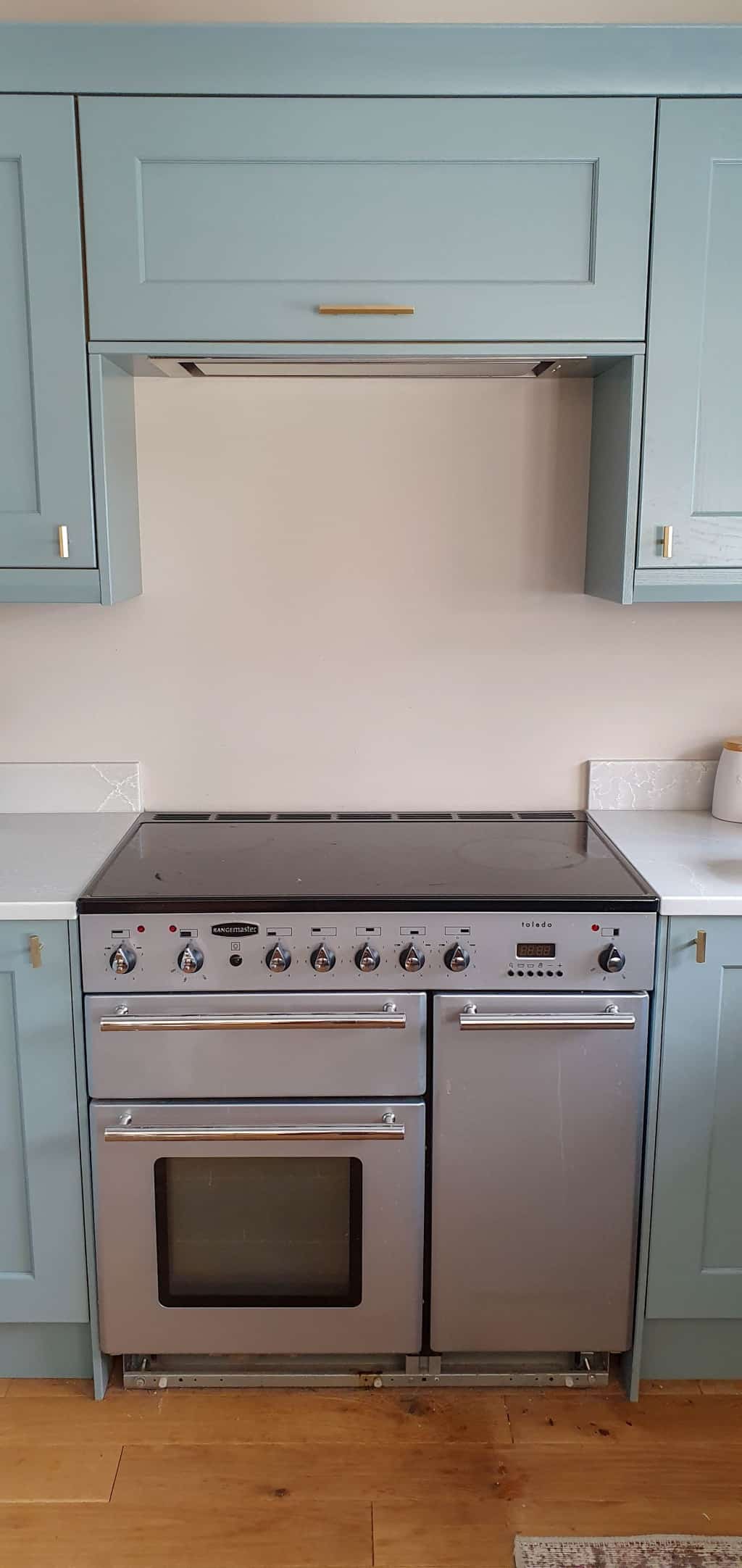 A modern kitchen features a silver freestanding range cooker with multiple knobs and two oven doors, set within light blue cupboards, white worktops, and metallic wall tiles for a sleek finishing touch.