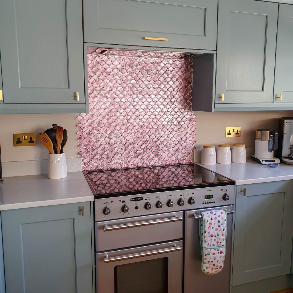 A modern kitchen with pale green cupboards, a large silver cooker, and a pink Vitreum Rosa tile splashback. Utensils and containers are on the white worktop, and a floral tea towel hangs from the cooker handle.