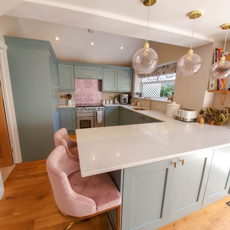 Modern kitchen with light blue cupboards, marble worktops, and wooden floor features three pink velvet stools at an L-shaped island. Glass pendant lights hang above, while Fired Earth Metallic Vitreum Rosa wall tiles create a striking pink splashback behind the cooker.