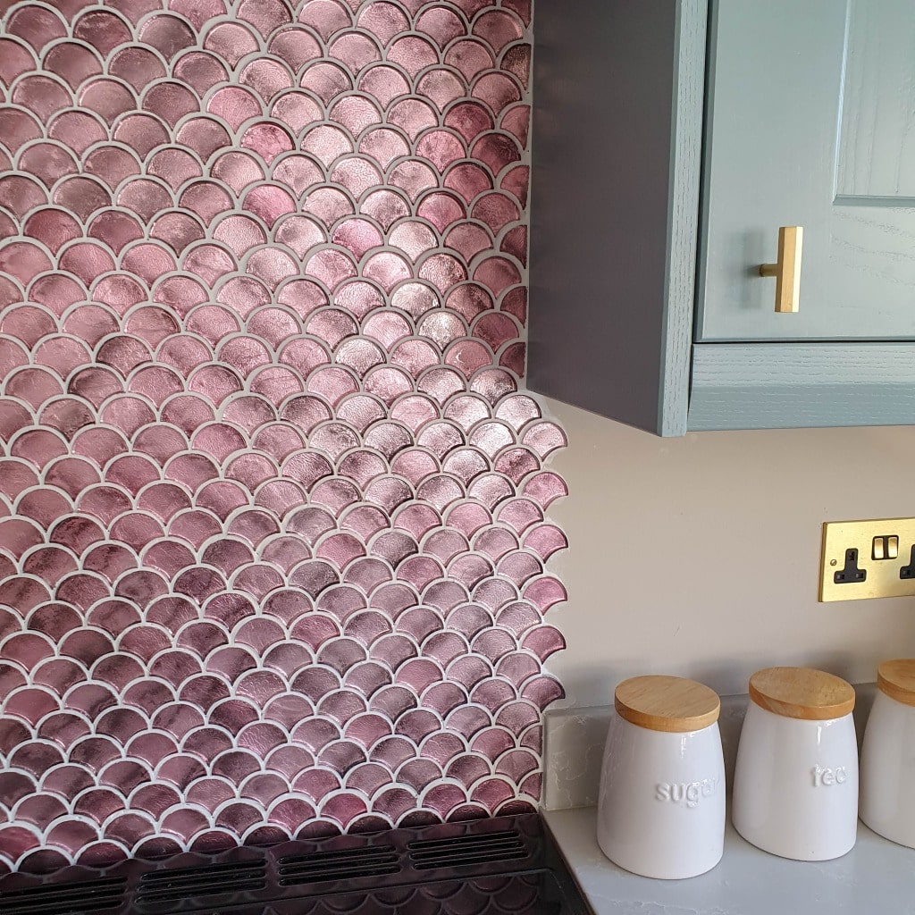 A kitchen splashback with shiny pink fan wall tiles—Metallic Vitreum Rosa by Fired Earth—next to a light grey cupboard, gold handle, electrical socket, and three white canisters labelled sugar, tea, and coffee with wooden lids.