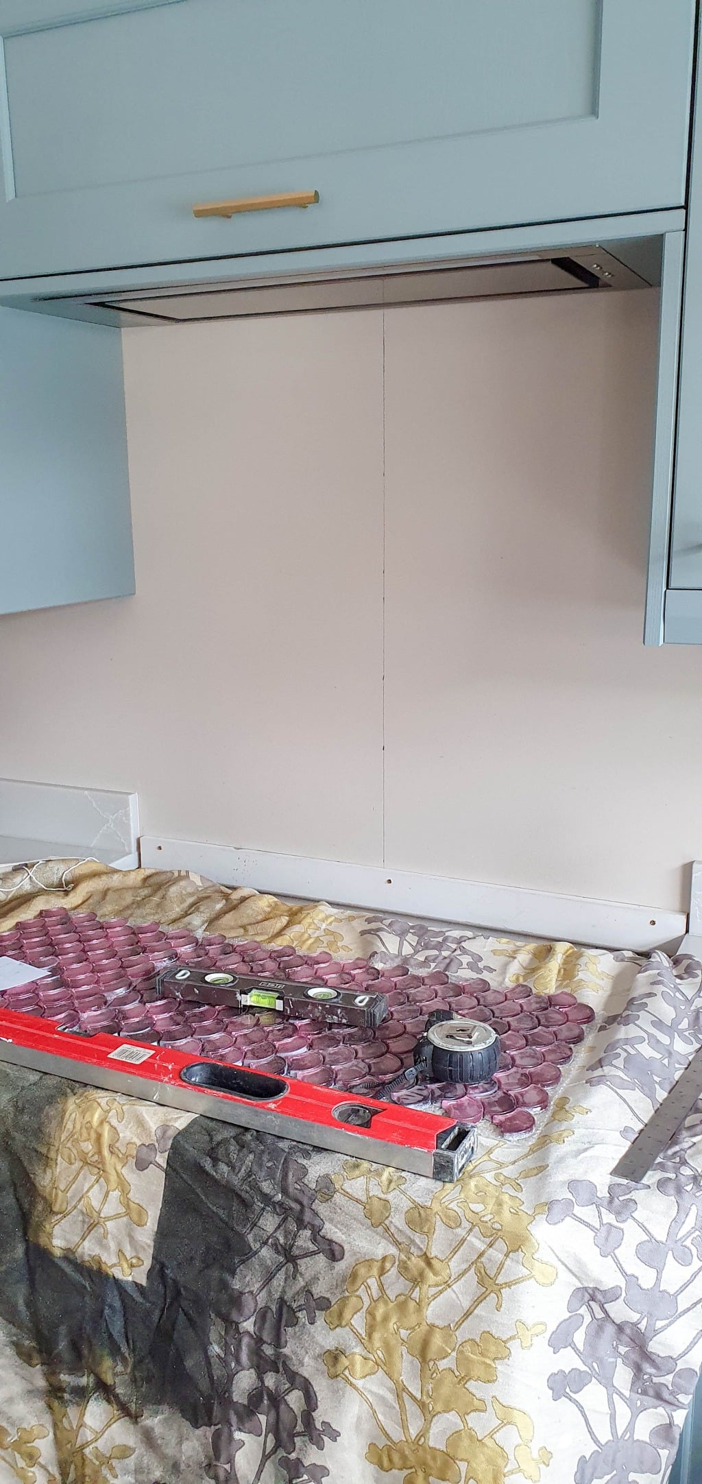A kitchen wall with light blue cupboards, a blank beige splashback area ready for wall tiles such as Metallic Vitreum Rosa from Fired Earth, and a spirit level and tape measure on a cloth-covered work surface. The wall appears to be in progress for renovation or installation.