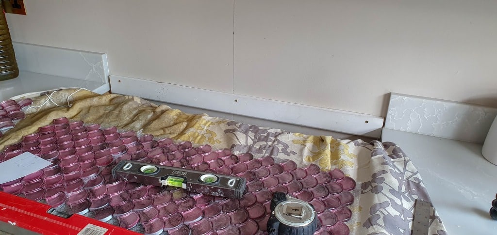 A kitchen worktop covered with a floral cloth and pink round Vitreum Rosa mosaic tiles, along with a spirit level, tape measure, and a red tool. The splashback area appears to be undergoing renovation.