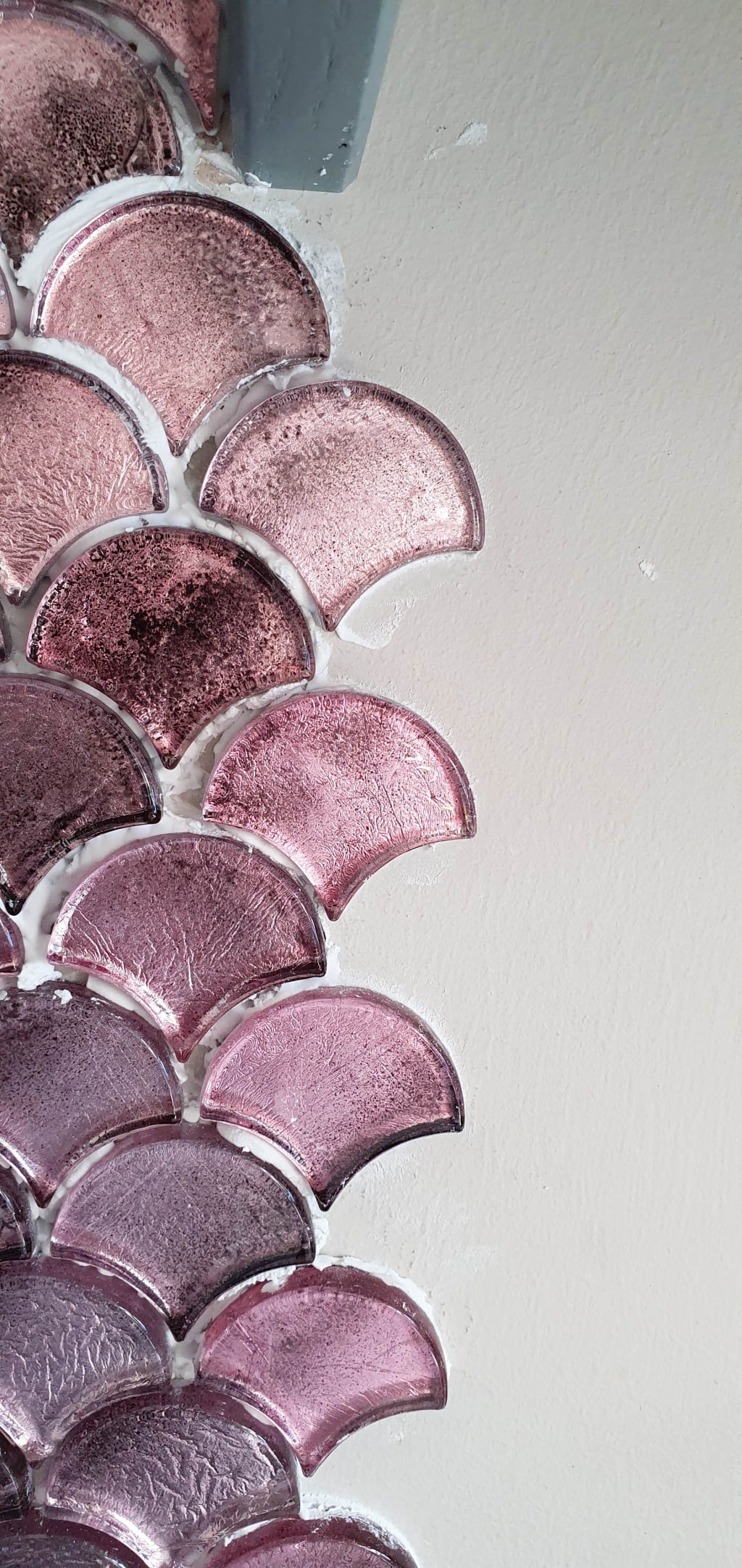 Textured, pink Vitreum Rosa tiles arranged in a fish scale pattern on a wall, with the left side tiled and the right side a plain, off-white surface.
