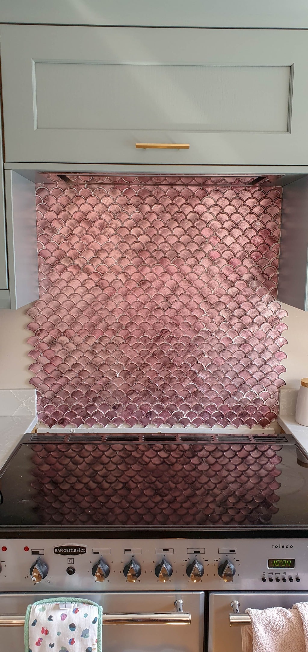 A kitchen splashback with pink Vitreum Rosa Tiles in a fish-scale pattern sits behind a black hob and oven, beneath pale green cupboards with a gold handle. A small utensil holder is on the right.