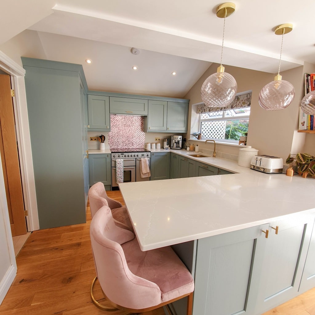 Modern kitchen with pastel green cupboards, marble worktop, wooden floor, pink velvet bar stools, and pendant lights. Fired Earth wall tiles add elegance behind the cooker, toaster, and coffee maker. Window offers a lovely garden view.