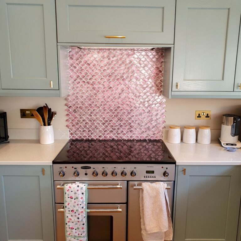 A modern kitchen with pale green cabinets, a stainless steel oven, light worktops, and Fired Earth Metallic Vitreum Rosa wall tiles in a pink fish scale pattern. Utensils, jars, a tea towel, and coffee machine sit neatly on the counter.