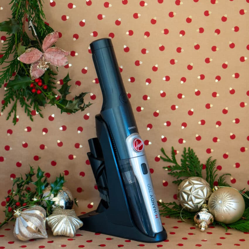 A handheld vacuum in a charging stand is displayed against festive, polka-dotted wrapping paper, surrounded by holiday decorations—an ideal home tech Christmas present featured in this season’s gift guide.