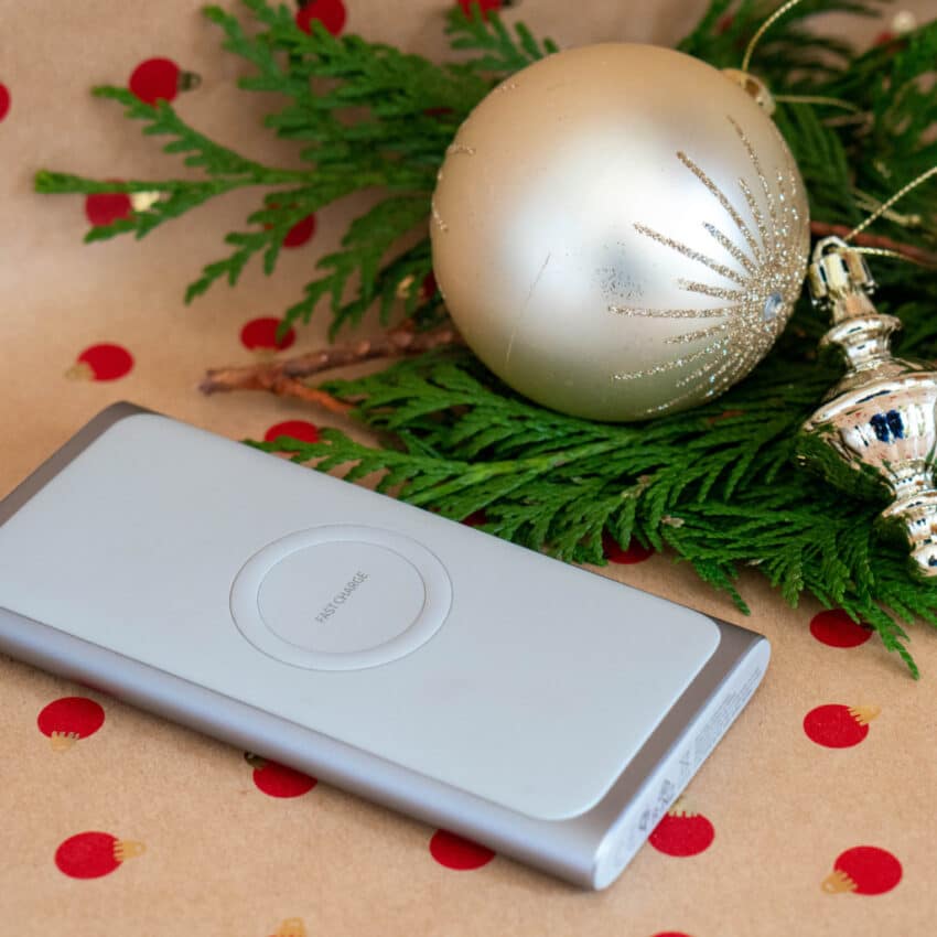 A silver wireless charging pad, perfect for any Home Tech lover, sits on festive wrapping paper with red dots, next to green pine branches, a gold Christmas bauble, and a silver bauble—an ideal Christmas present.