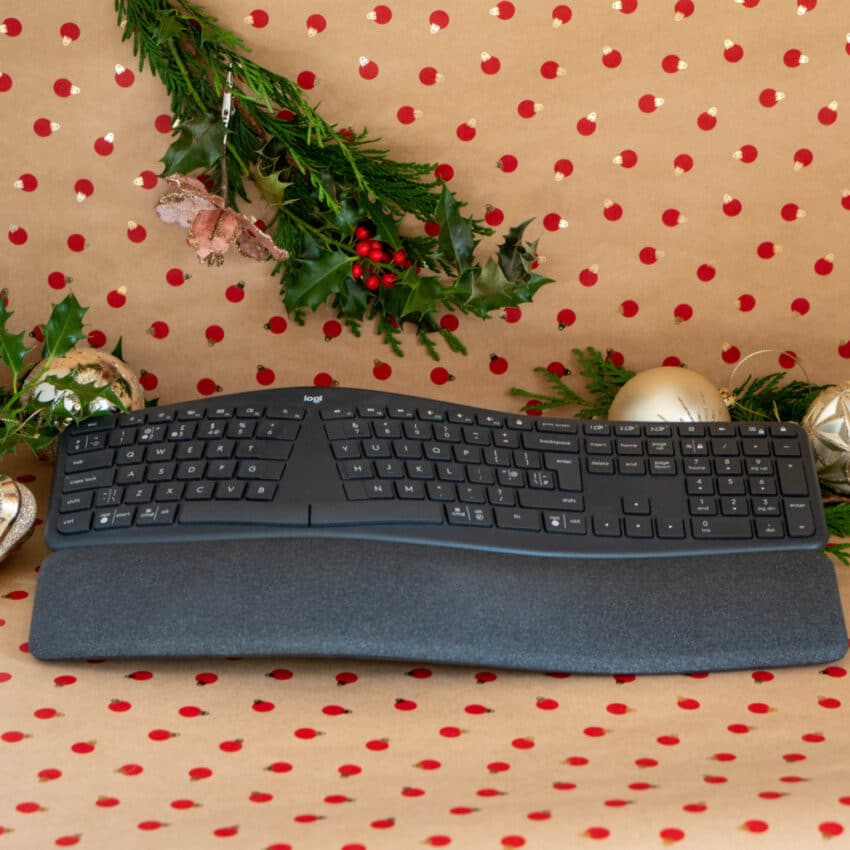 A black ergonomic keyboard with a wrist rest sits on festive wrapping paper with red dots, surrounded by ornaments and greenery—a perfect addition to any Home Tech Christmas Gift guide.