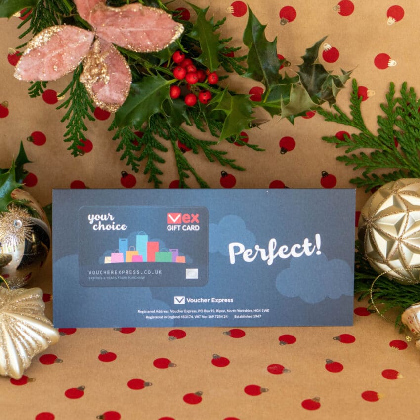 A VEX gift card rests on festive, red spotty wrapping paper, surrounded by gold baubles, holly, pine branches, and pink flowers—a perfect pick for tech gifts in any Christmas Gift Guide.