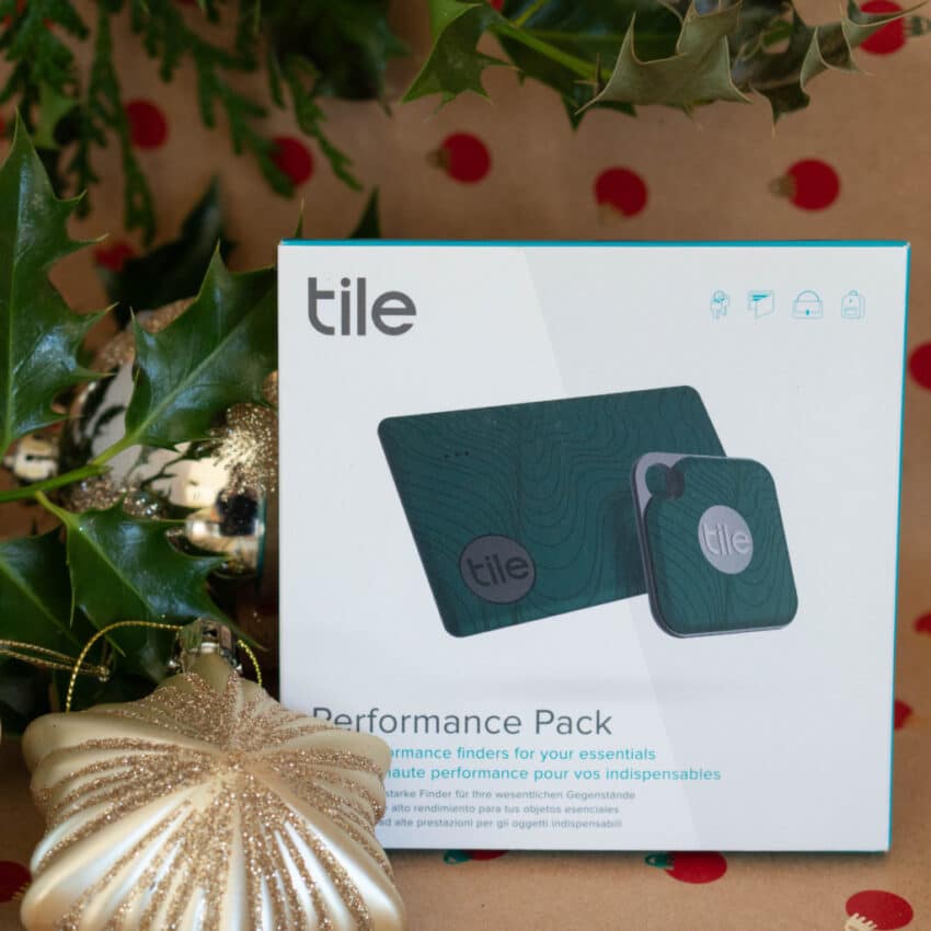 A Tile Performance Pack box sits among green holly leaves, a gold ornament, and festive decorations on brown paper with red dots—a perfect addition to any Christmas Gift Guide for top home tech or tech gifts. The box displays two Tile Bluetooth trackers.