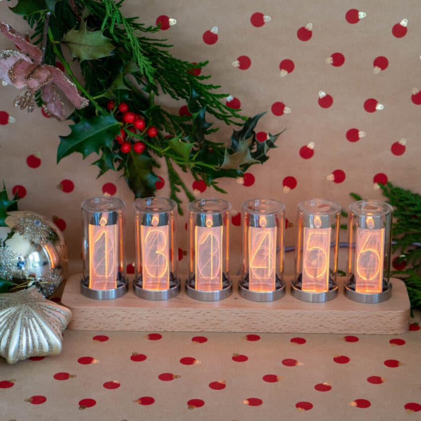 A Nixie tube clock displays 13:04:56 on a wooden base, surrounded by Christmas decorations—holly, baubles, and festive red-spot wrapping paper—making it a unique Home Tech choice for your Christmas Gift list.