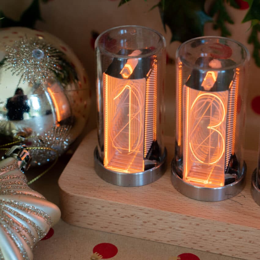 Two glowing Nixie tubes display the numbers 1 and 3 on a wooden base. Surrounded by festive ornaments, this unique piece is a standout in any Home Tech collection and makes a perfect addition to your Christmas Gift list.
