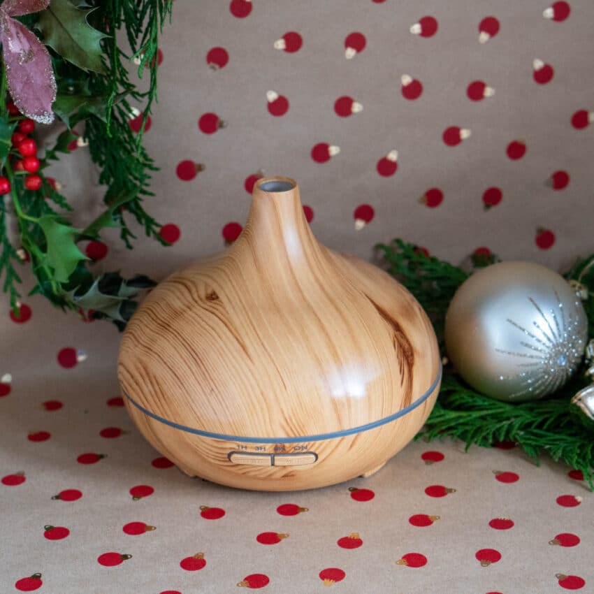 A wooden-look essential oil diffuser sits on a festive, red and tan polka dot background, surrounded by green holiday foliage, a gold bauble, and a decorative ribbon—a perfect addition to any Christmas gift or home tech gift guide.