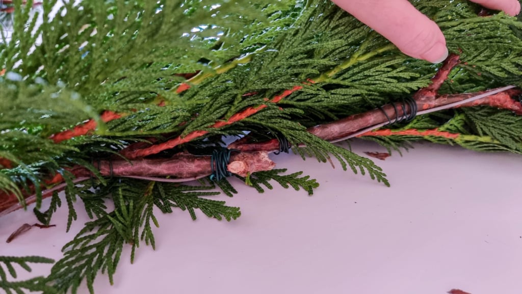 A hand demonstrates how to make a real Christmas garland, arranging green evergreen branches and securing them together with dark wire on a white surface.
