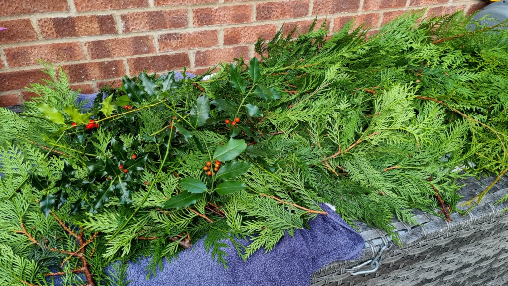 A pile of freshly cut evergreen branches, perfect to make Christmas garland, includes holly with red berries and green foliage, laid on a purple towel over a wicker surface against a brick wall.