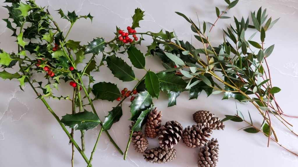 Three sprigs of holly with red berries, a eucalyptus branch, and several small pinecones are arranged on a white marble surface—perfect inspiration if you're learning how to make a real Christmas garland.