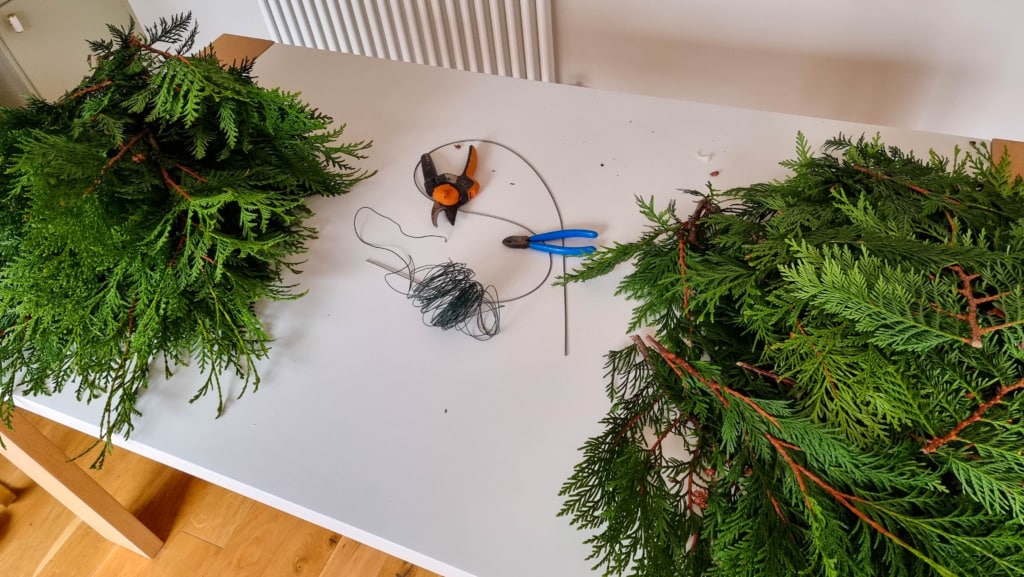 A white table with green tree branches, wire, pliers, scissors, and a spool of string suggests a Real Christmas Garland project. Indoors with wooden flooring and a radiator in the background, it's perfect for learning How To Make Christmas Garland.