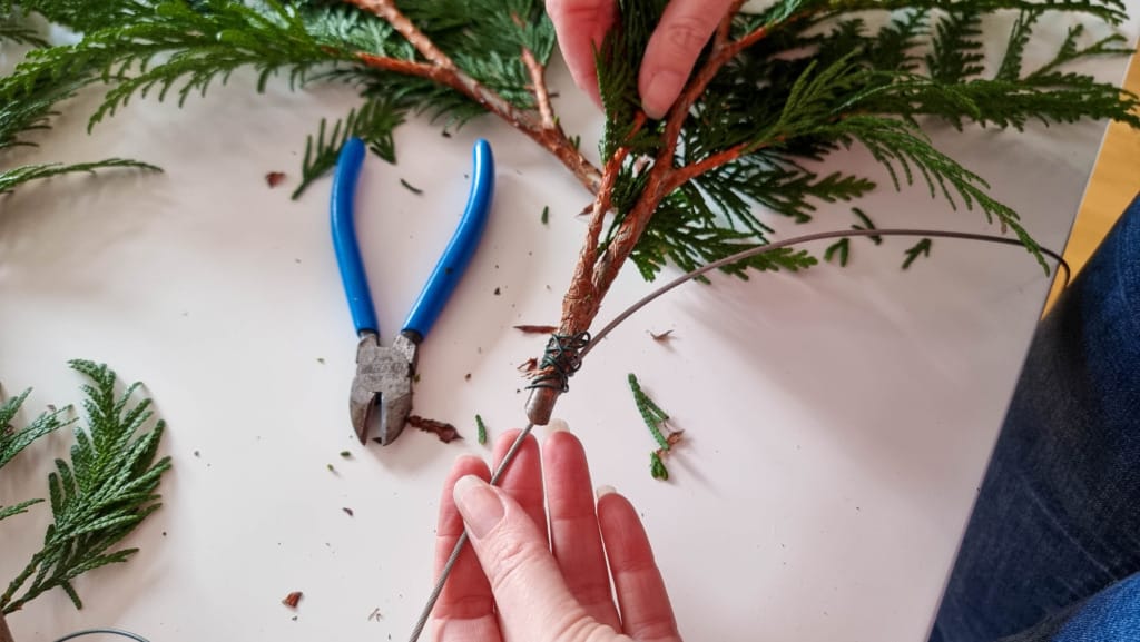 A person’s hands twist florists’ wire around evergreen branches to make Christmas garland on a white surface. Blue wire cutters and scattered greenery are visible nearby.