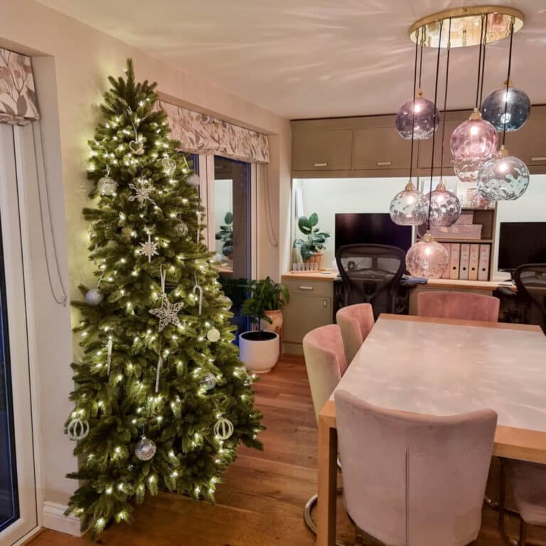 A tall, narrow Chelsea Flatback Christmas Tree decorated with white lights and silver baubles stands in the corner of a modern dining room with pink chairs, a white table, hanging glass fittings, and a computer desk.