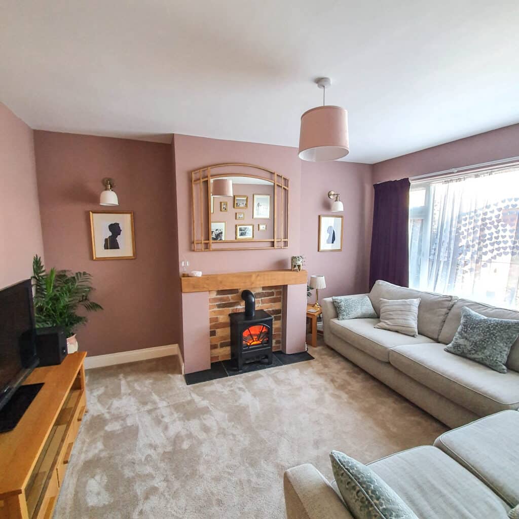 A cosy living room with pink walls, beige carpet, a light grey sectional sofa, and a wooden mantelpiece fireplace showcases how you can turn your house into a home with thoughtful touches like framed artwork, TV setup, and sheer-curtained windows.