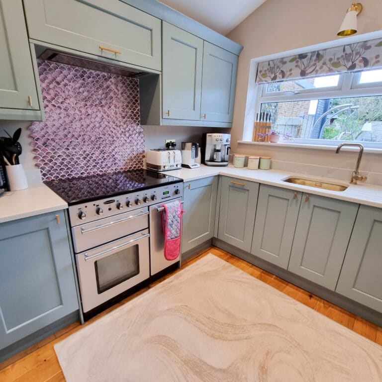 A modern kitchen with light blue cupboards, a white cooker, pink patterned splashback, countertop appliances, a sink under a window with floral valance, and a beige washable rug—perfect for easy cleaning and inspired by my personal review.