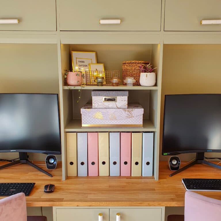 A tidy home office setup with two monitors, keyboards, pastel-coloured file binders, decorative boxes, plants, and framed prints arranged on wooden shelves with a light green built-in desk showcases smart home organisation.