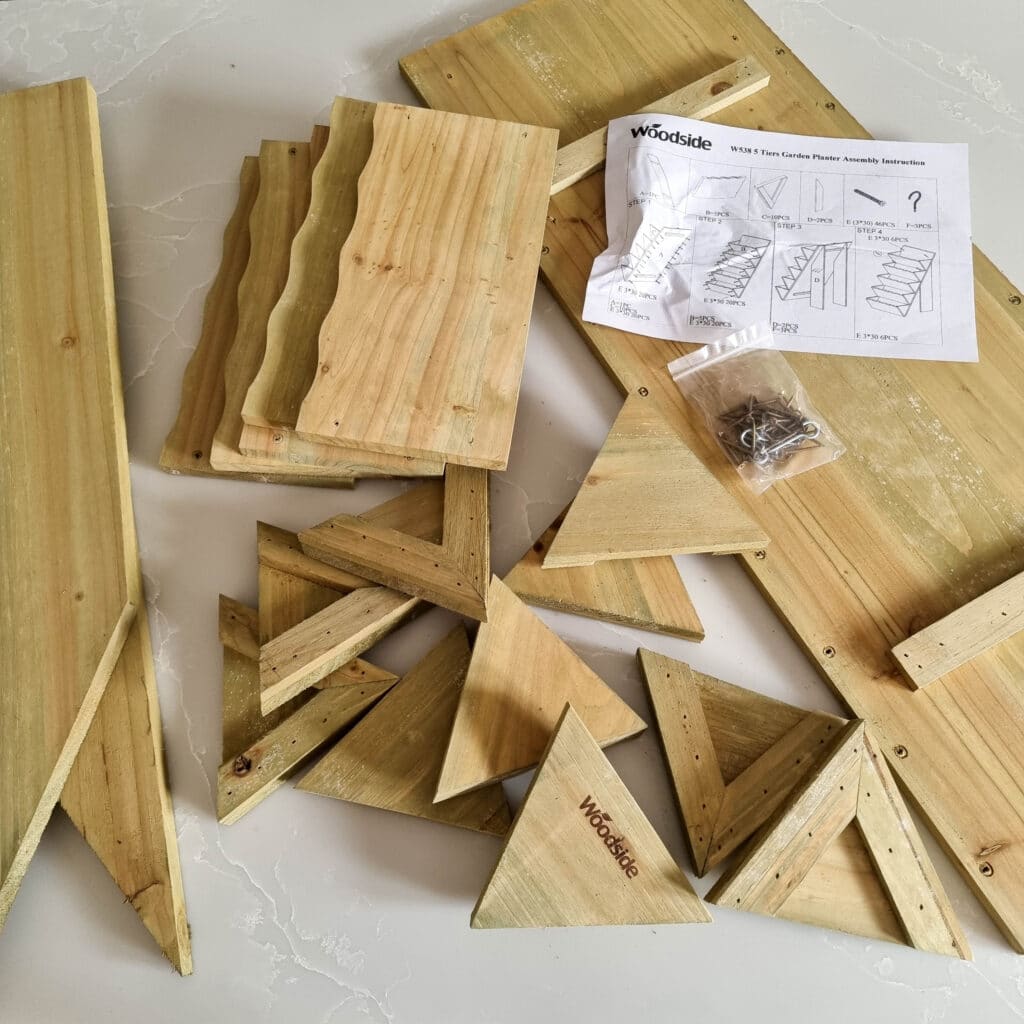 Several wooden planks and triangular pieces, screws in a plastic bag, and an instruction sheet are laid out on a white surface, ready for assembly into a Wooden Herb Planter. Some wood pieces are branded with Woodside.
