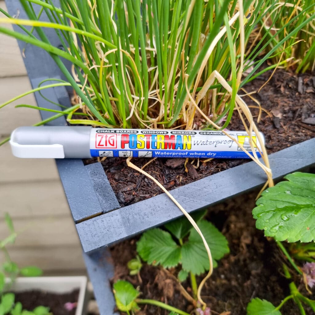 A silver ZIG Posterman waterproof marker rests on the edge of a wooden herb planter filled with green chives and other plants, with leaves and soil visible below.