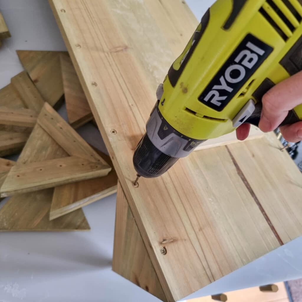 A person uses a yellow Ryobi power drill to drive a screw into a wooden board, assembling what appears to be a wooden herb planter, with additional wooden pieces and triangular sections visible in the background.