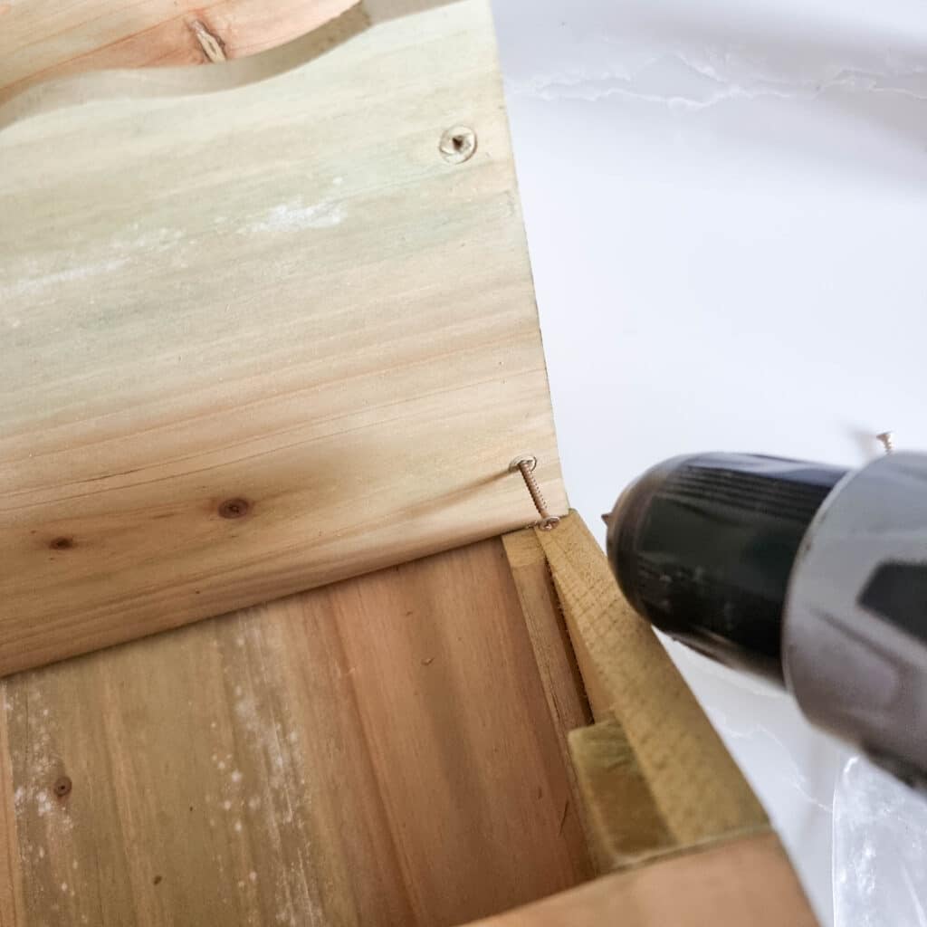 A close-up of a person using a power drill to drive a screw into the corner of a wooden herb planter, securing two pieces of light-coloured wood together.