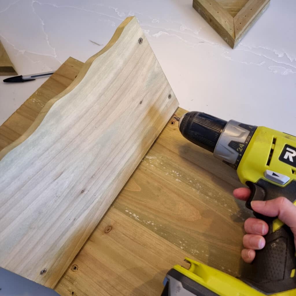 A person uses a Ryobi power drill to attach a piece of light-coloured wood to another board, assembling a wooden herb planter on a white surface with a black pen nearby.