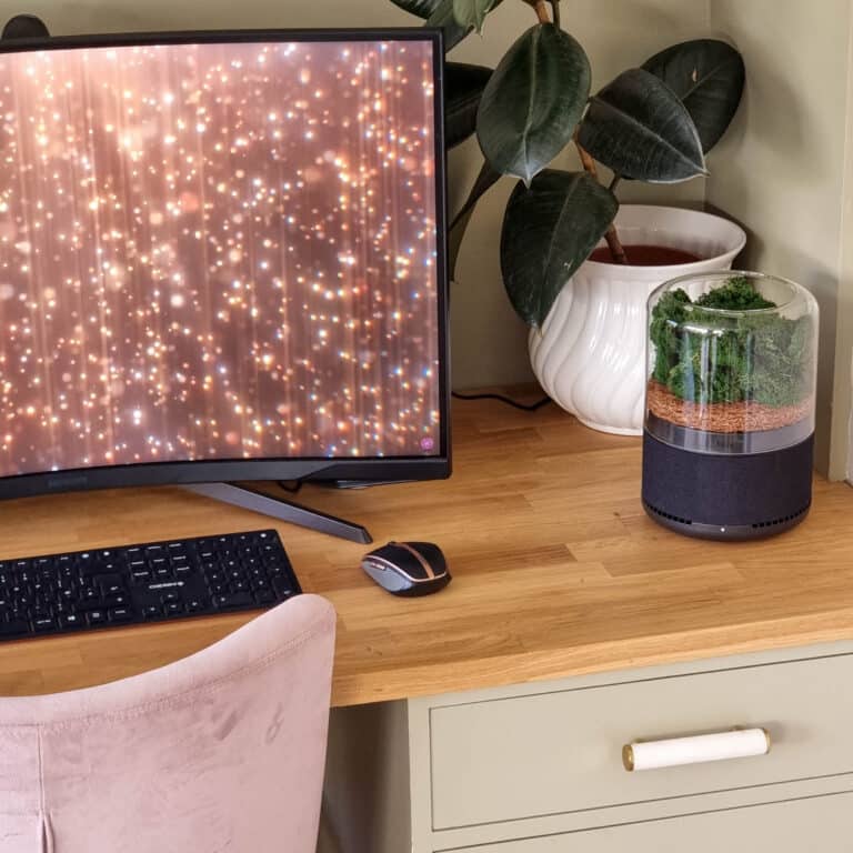A wooden desk with a computer monitor showing a sparkly light pattern, keyboard, mouse, potted plant, small terrarium, and even a Briiv Air Purifier. A soft pink chair and green drawers add to the inviting workspace ambience.